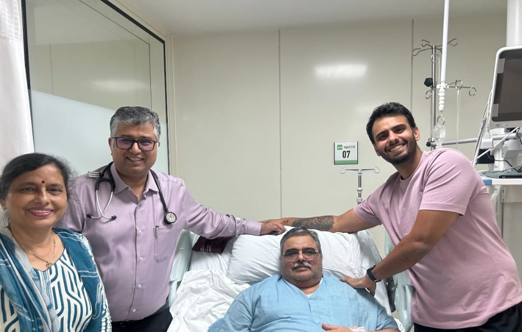 Vidit Aren and his mother standing beside his father in the hospital bed with Dr. Manish Aggarwal at Yashoda Medicity